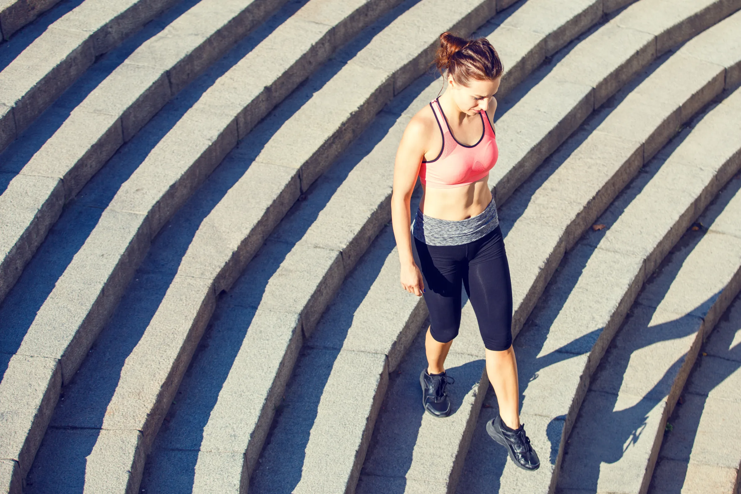 A slim woman in a pink top and black capris descends stone stairs.