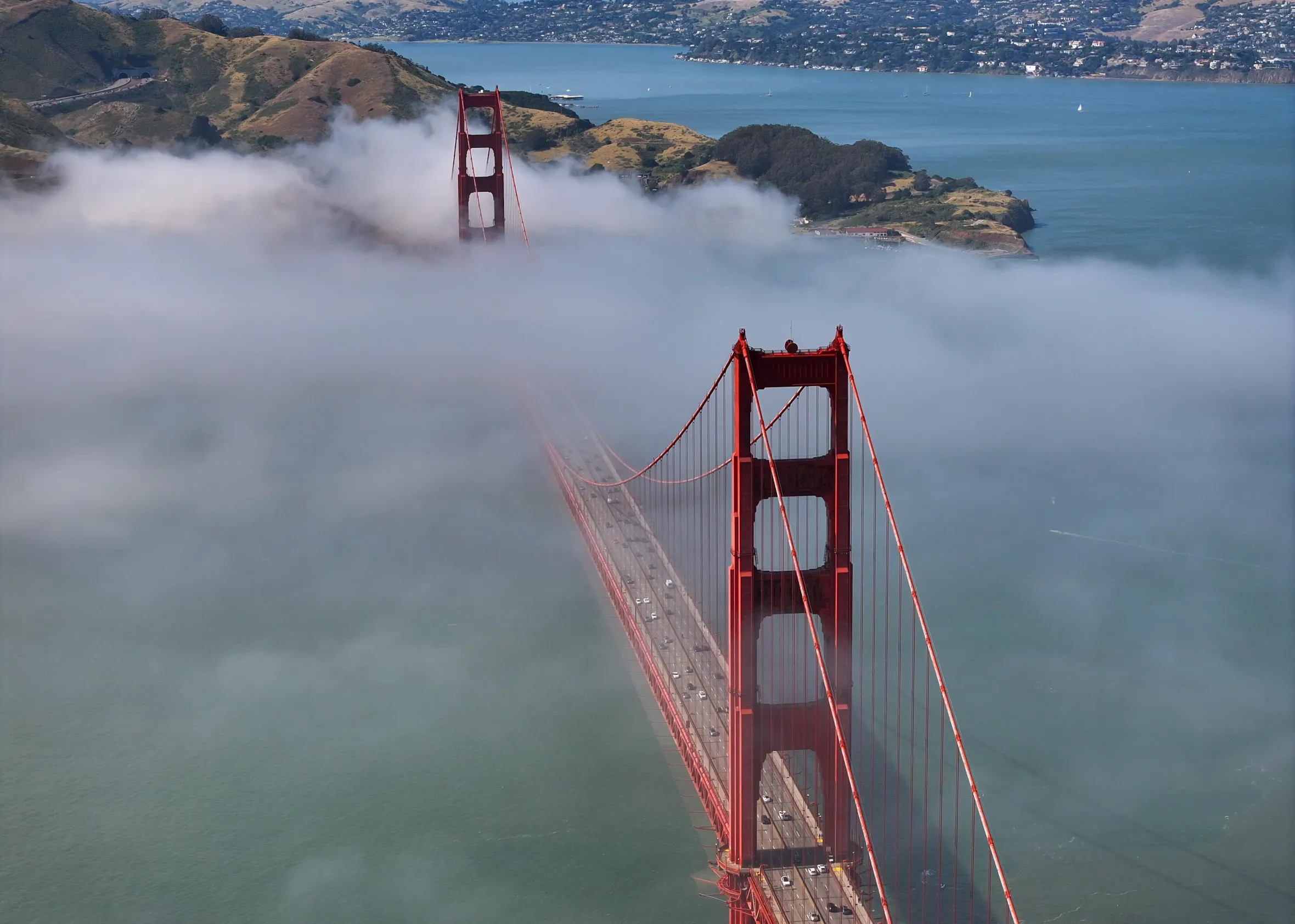 Aerial view of the Golden Gate Bridge partially obscured by thick fog, with the distant San Francisco Bay visible.
