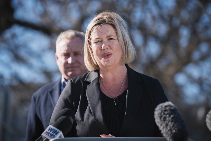 A female politician speaks from a podium with a man standing behind her