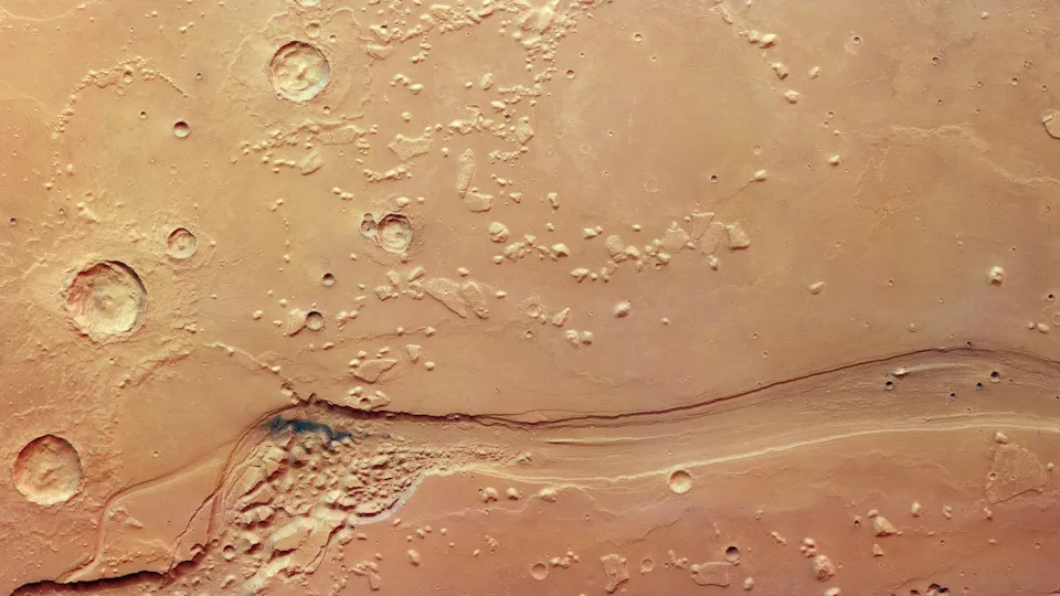 Looking down on a patch of Mars's surface, with a winding canyon carving from the left to the right of the frame. On the left, the canyon widens and is filled with lumpy, knobbly terrain. On the right, the canyon is more straight and streamlined. Surrounding the canyon is more lumpy terrain, but on top of a smoother surface