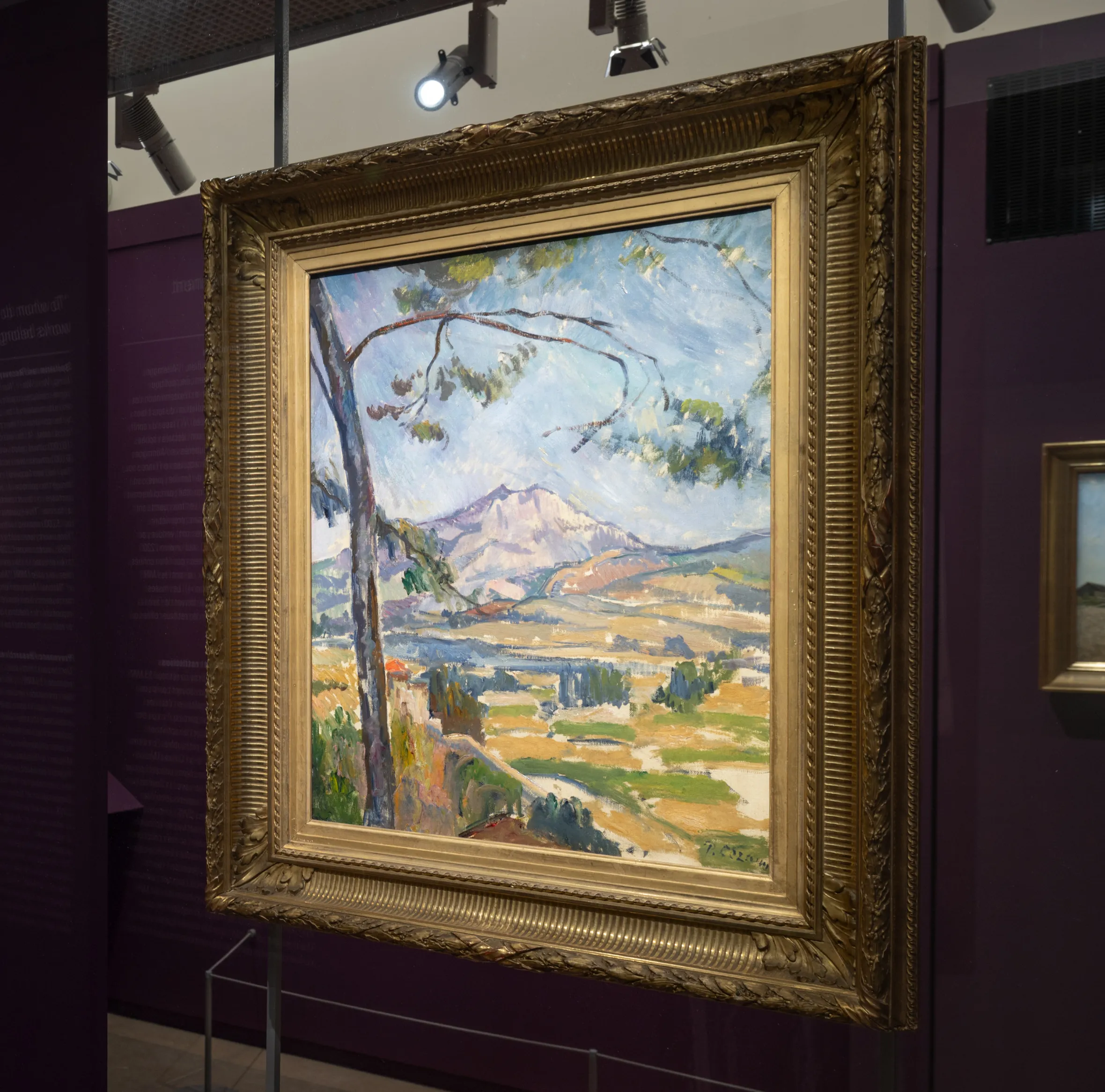 Painting of a mountain landscape with trees, houses, and fields, displayed in a museum gallery with purple walls.