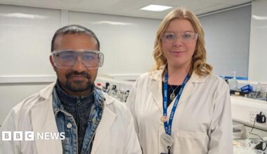 A man stands on the left wearing a white lab coat over a denim jacket with safety goggles over his glasses. He has short dark hair and dark facial hair. To his right, a woman stands wearing a white lab coat done up with a blue staff lanyard and necklace on. She has shoulder length curled blonde hair, behind then is a lab desk.