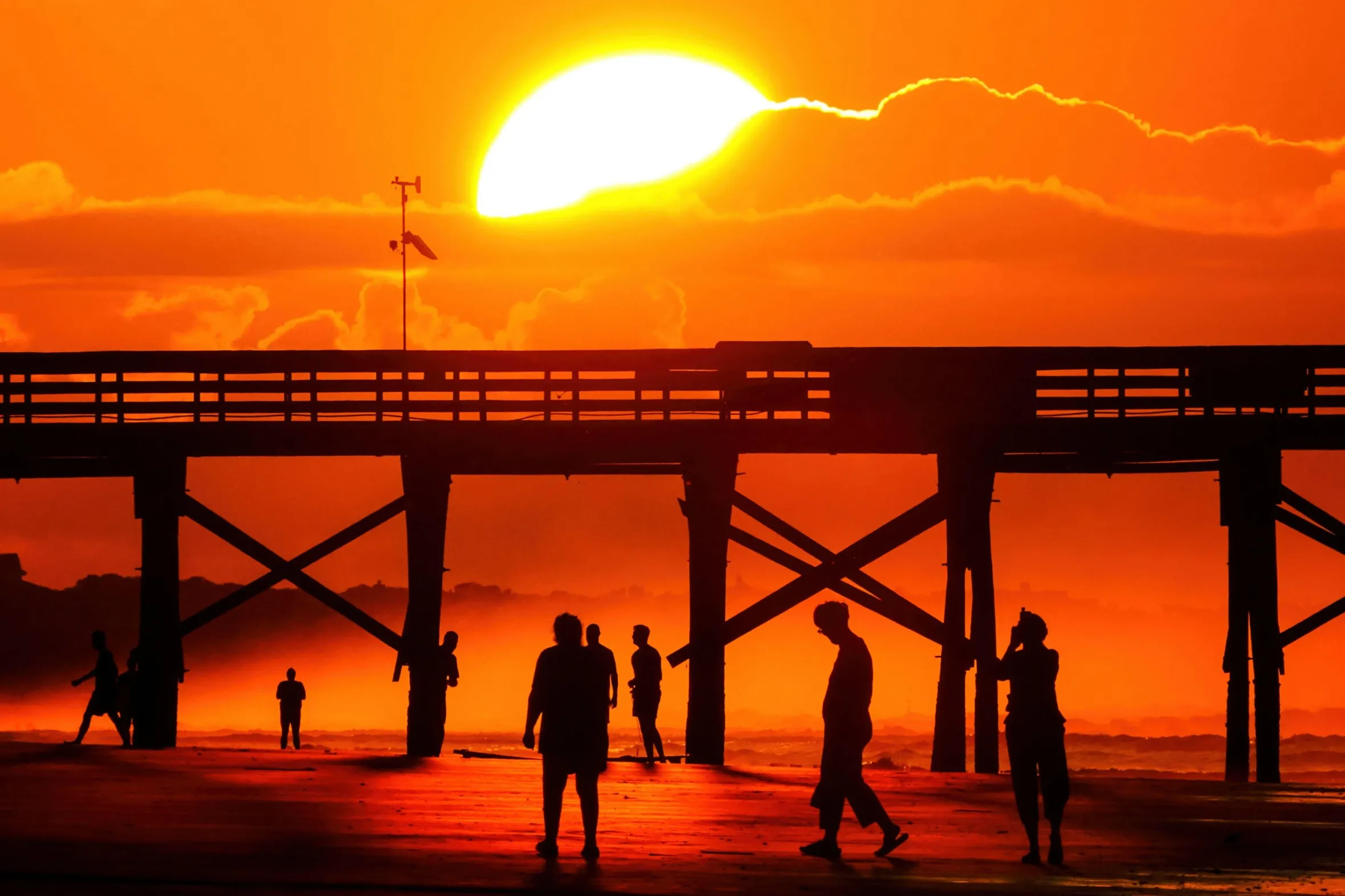 Silhouettes of beachgoers walking along a beach near a fishing pier at sunrise.