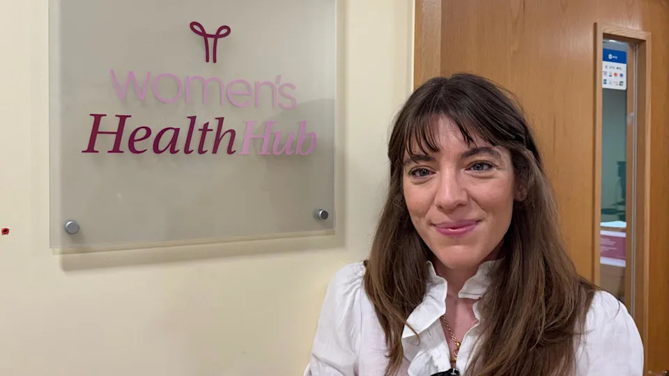 Dr Jessica Langtree-Marsh is smiling at the camera in a heads-and-shoulders photo. She has long brown hair and is wearing a white top. She is standing next to a sign in a corridor that says "Women's Health Hub". There is a door behind her.