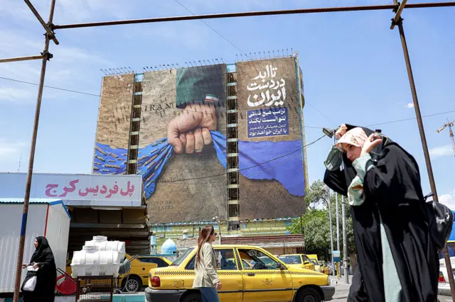 A woman adjusts her headdress as she walks near a billboard on the facade of a building depicting the Strait of Hormuz with a caption in Persian reading "Forever in Iran's Hand"