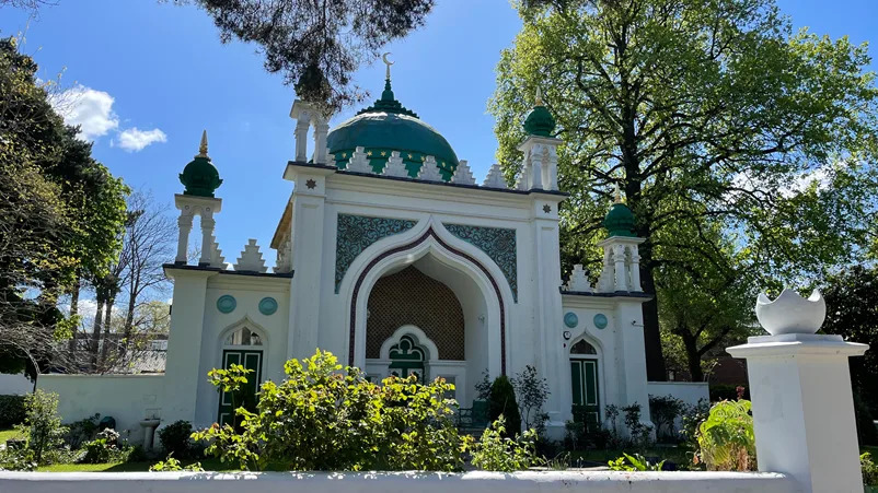 A white and green domed mosque stands in bright sunshine under a blue sky. There are ornate decorations and tiling on the mosque with trees and shrubs around the outside.