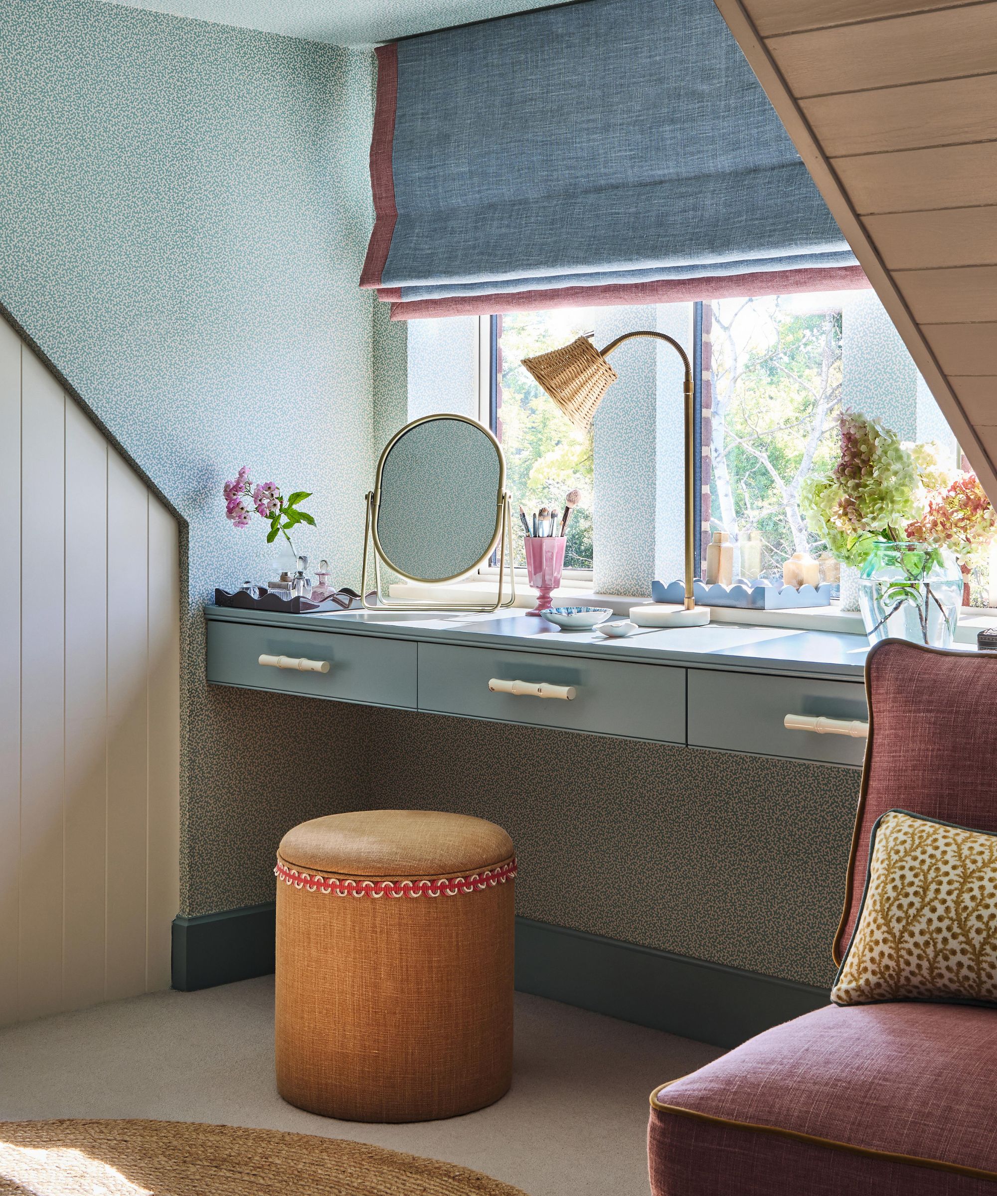 the corner of an attic master bedroom with green ditsy print wallpaper, a desk built under the window, small stool, blue blind and a slipper chair