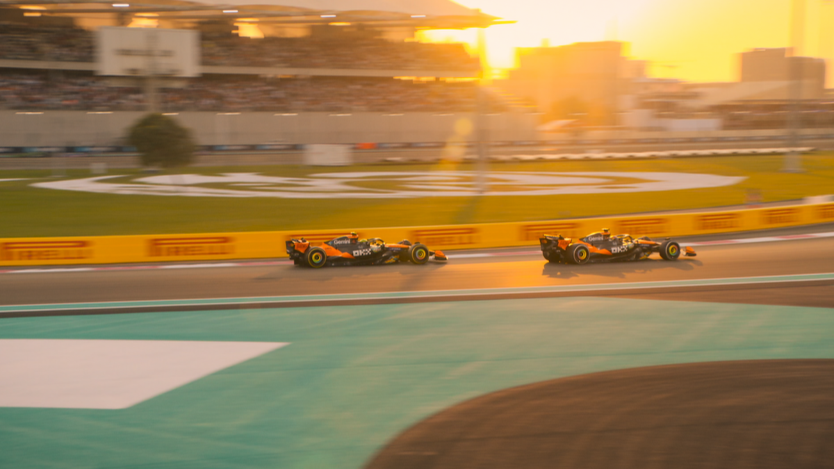 Two Formula 1 cars racing on a circuit at sunset, with stadium seating filled with spectators in the background and colorful branding visible along the track barriers.