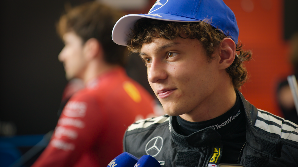 Young race car driver in a Mercedes team suit and blue cap speaking to the media in a press area, with microphones visible and another person in a red racing suit blurred in the background.