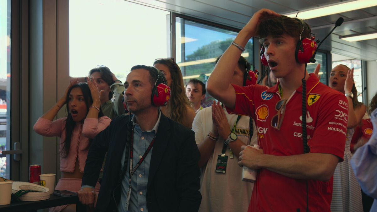 A group of people in a Formula 1 team suite react with shock and surprise, wearing headsets, watching an event through the windows, with a race team member in Ferrari gear in the foreground.