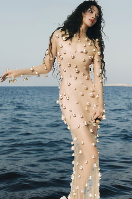 A woman stands by the sea wearing a sheer, nude-colored dress adorned with large pearl-like beads. She has long, wavy dark hair and poses with one arm slightly raised, looking confidently toward the camera.