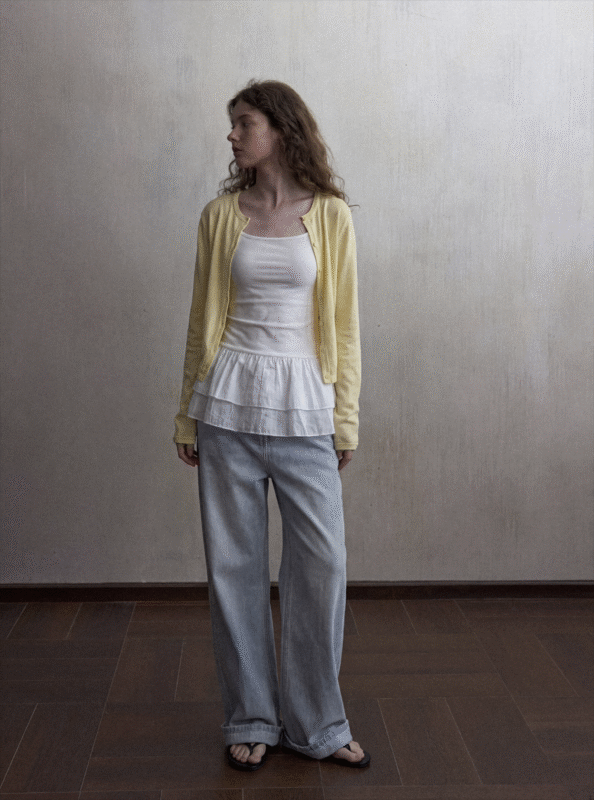 A woman with wavy hair stands indoors wearing a yellow cardigan over a white top with a ruffled hem, loose light blue jeans, and black sandals, looking to her left against a plain light-colored wall.