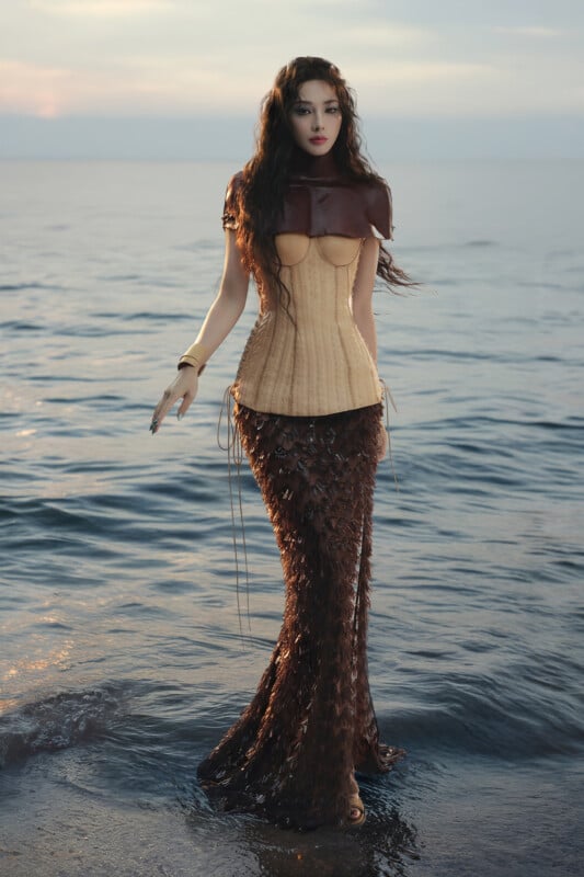 A woman stands at the edge of the sea during sunset, wearing a unique outfit with a structured beige corset, dark shoulder piece, and a long, textured, scale-like brown skirt that resembles a mermaid tail.