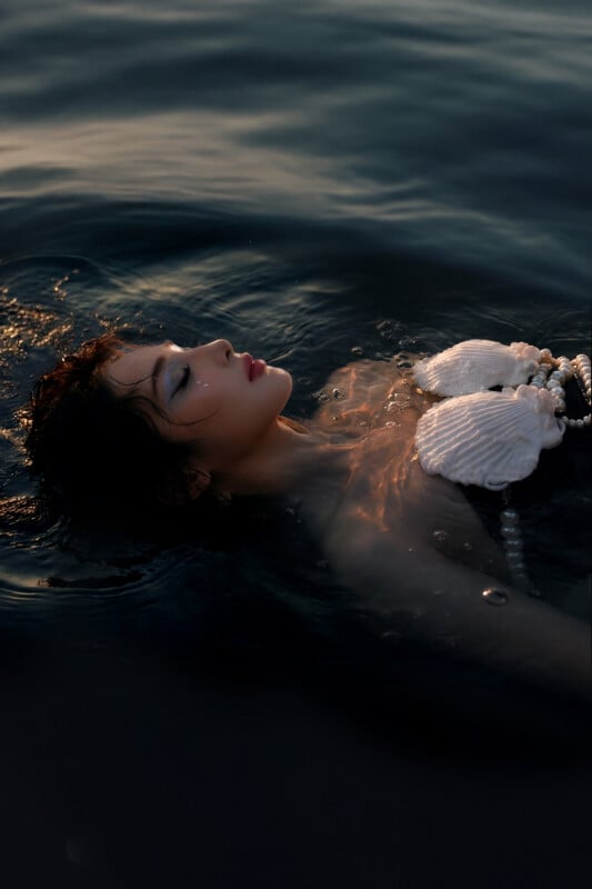 A person with closed eyes floats peacefully in dark water, wearing a seashell top and a pearl necklace, with soft light reflecting off the water’s surface.