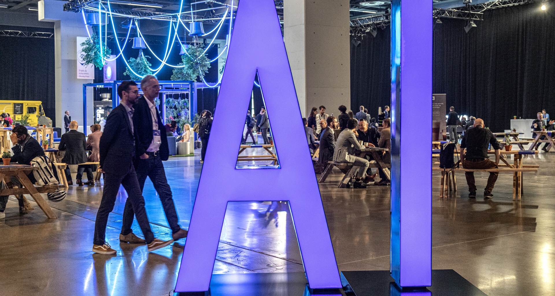 Large purple letters spell out “AI” on a display at the All In AI conference in Montreal. Two men wearing blazers walk by on the left, while other attendees sit at tables and tree-like decorations hang from the ceiling in the background.