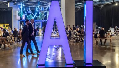 Large purple letters spell out “AI” on a display at the All In AI conference in Montreal. Two men wearing blazers walk by on the left, while other attendees sit at tables and tree-like decorations hang from the ceiling in the background.