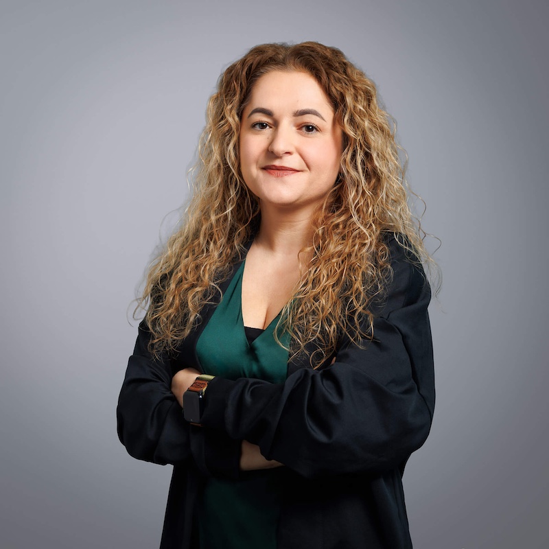 Young woman with long, curly dark-gold hair standing with her arms folded.