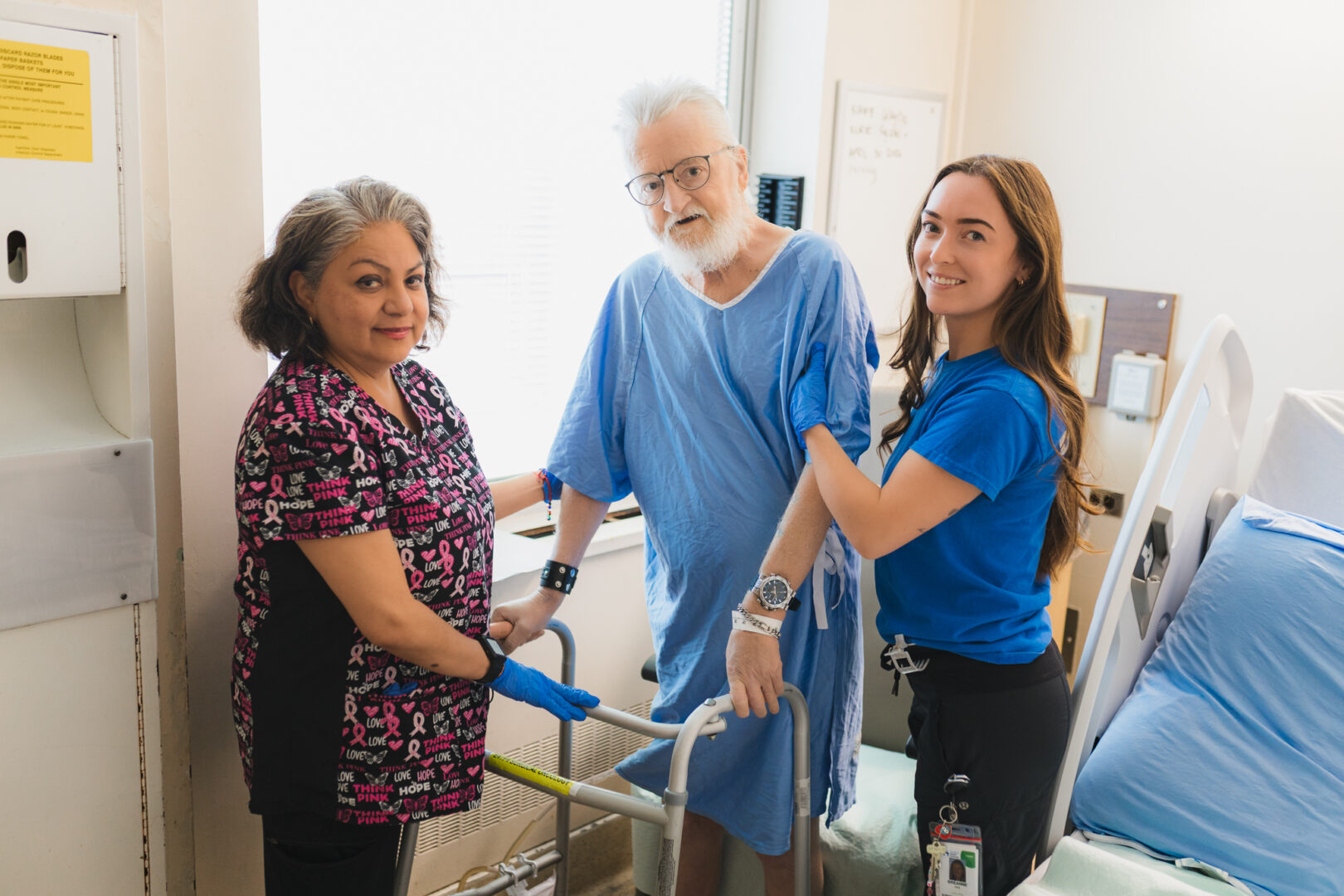 A hospital patient in a blue gown holds his walker beside his bed while two health-care workers support him. They are all looking at the camera.