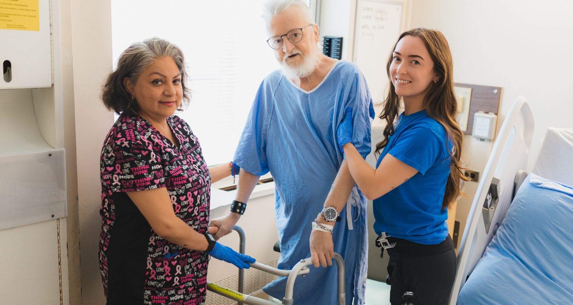 A hospital patient in a blue gown holds his walker beside his bed while two health-care workers support him. They are all looking at the camera.