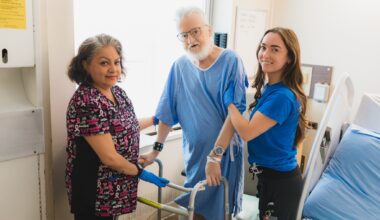 A hospital patient in a blue gown holds his walker beside his bed while two health-care workers support him. They are all looking at the camera.