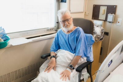 A hospital patient sits in a hospital chair with a blanket on his lap.
