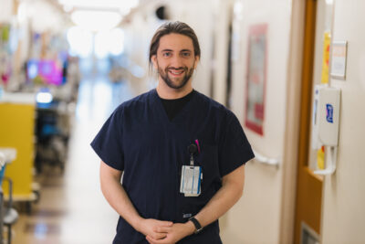 Headshot of nurse Cavan Stoddart, wearing dark blue scrubs. He has long hair and a beard.