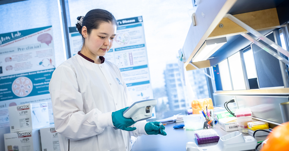 An individual in a lab coat and blue gloves looks at a device at a counter in lab setting.