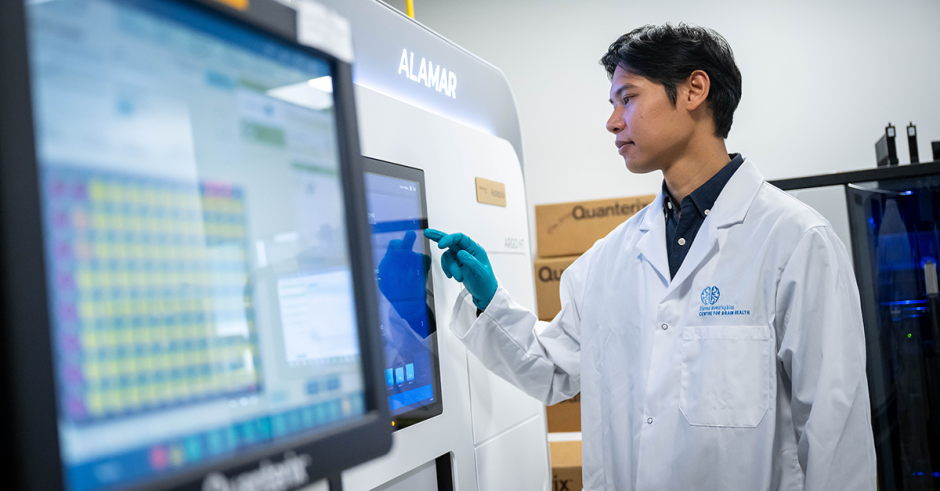 An individual in a lab coat stands on the right of a medical device as tall as they are. In the foreground there's a screen on the device. They individual uses the touch screen on the device with a gloved hand. 