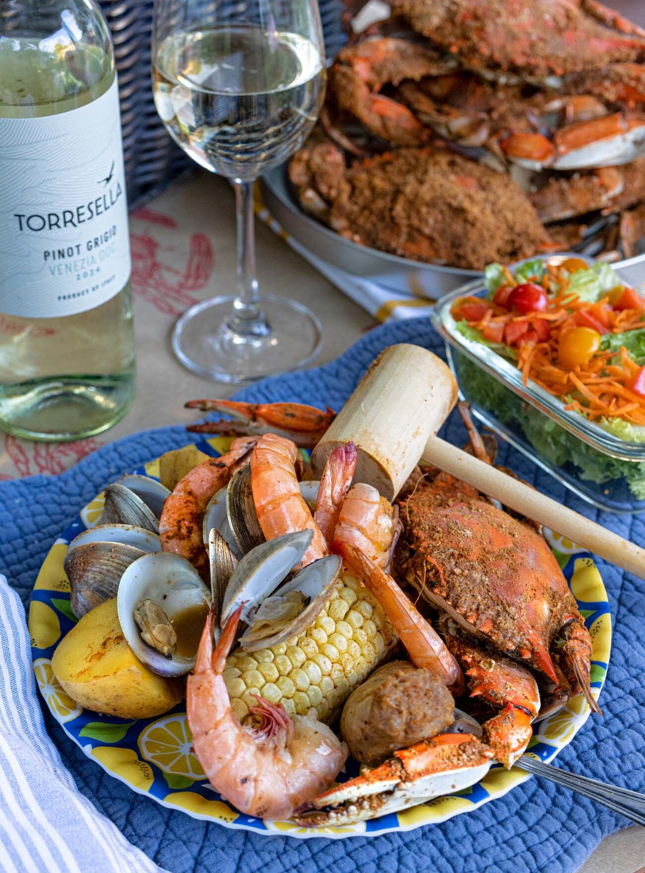  A Brampton 1860 Maryland crab boil dinner served on a decorative lemon-print blue and yellow plate on a blue quilted placemat, featuring a whole steamed blue crab seasoned with Old Bay spice, large shell-on shrimp, steamed clams, a half ear of corn, red potatoes, and a wooden crab mallet, alongside a bottle of Torresella Pinot Grigio Venezia 2024 and a poured glass of white wine, with a platter of additional steamed blue crabs and a colorful garden salad with cherry tomatoes visible in the background
