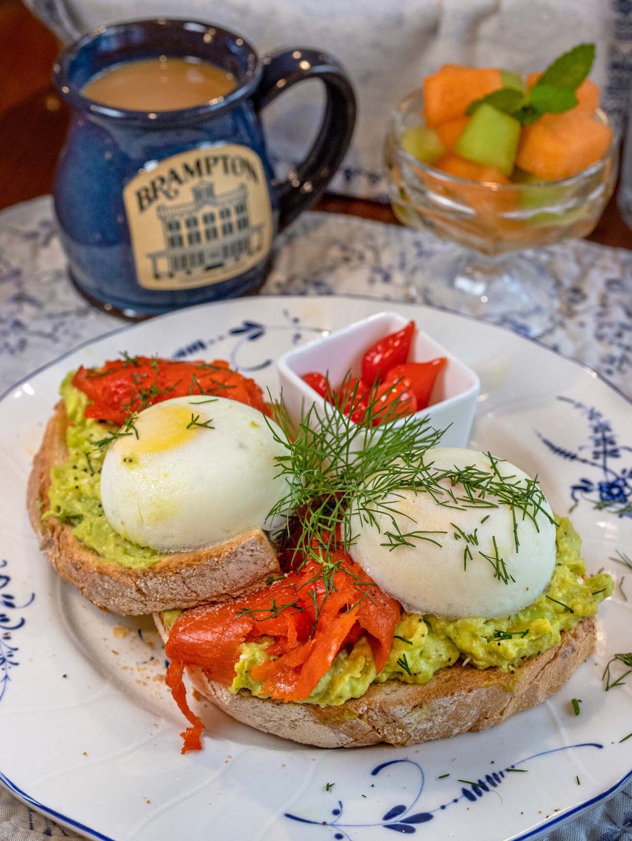 A close-up of a Brampton 1860 breakfast plate on blue and white china featuring two thick slices of artisan toast generously topped with smashed avocado, layers of smoked salmon, two perfectly poached eggs with set whites and soft yolks, fresh dill garnish, and a small ramekin of roasted cherry tomatoes on the side, with a blue Brampton branded coffee mug with cream, a basket of bread, and a fresh fruit cup with melon balls in the background