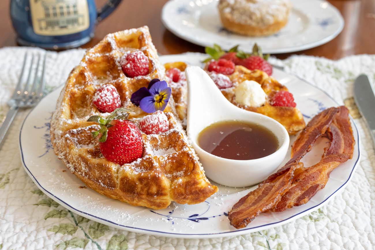 A Brampton 1860 breakfast plate on blue and white china featuring two golden Belgian-style waffles dusted with powdered sugar and topped with fresh strawberries, raspberries, and a purple and yellow edible viola flower, served with a white ceramic pouring vessel of maple syrup, a dollop of whipped cream, and three strips of crispy local bacon, with a blue Brampton branded coffee mug and a pastry on a side plate visible in the background on a white quilted tablecloth