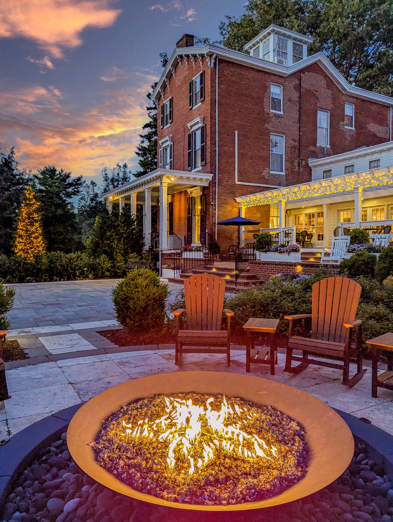The Brampton 1860 firepit burning bright at dusk with Adirondack chairs surrounding it and the illuminated historic red brick manor house glowing against a dramatic orange and purple spring sunset sky in Chestertown, Maryland