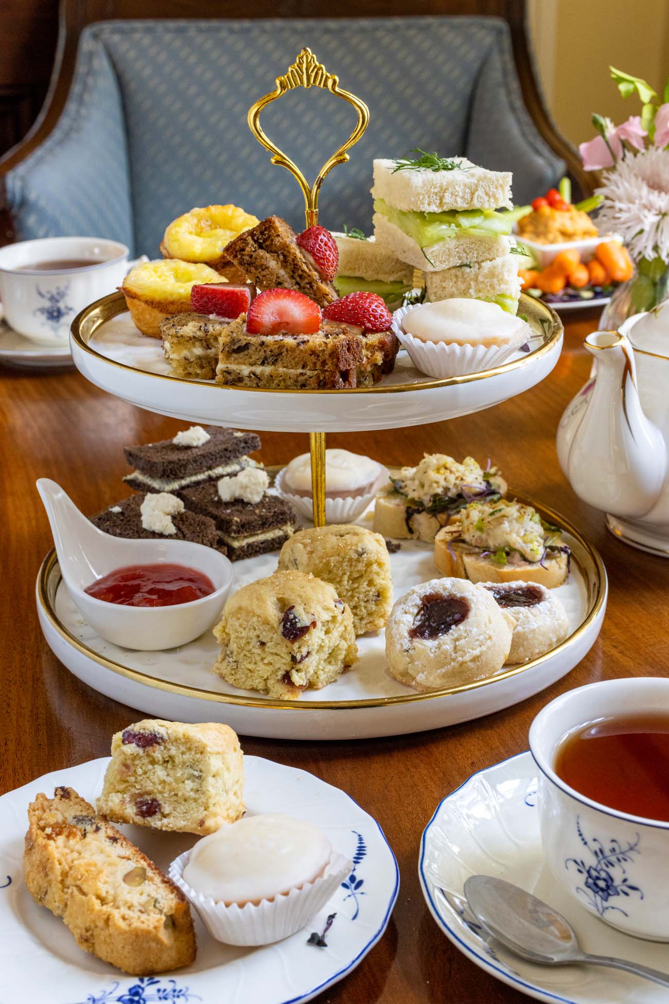 A two-tier gold-handled tea stand set on a wooden antique table at Brampton 1860, loaded with fresh-baked scones with jam, finger sandwiches, brownies, thumbprint cookies, lemon tartlets, and fruit bars topped with fresh strawberries, accompanied by a floral rose teapot and two cups of brewed tea on blue and white china