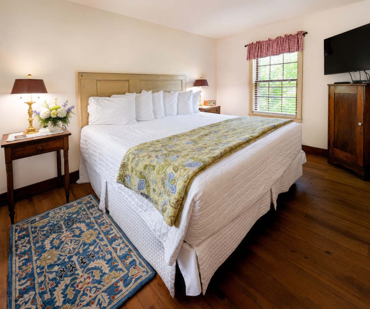 The king bedroom of the Fairy Hill Suite at Brampton 1860, featuring a white quilted bedspread with a floral runner, hardwood floors, a blue and cream Persian area rug, a dark wood armoire, brass table lamps, a small vase of fresh flowers, and a window overlooking green spring foliage