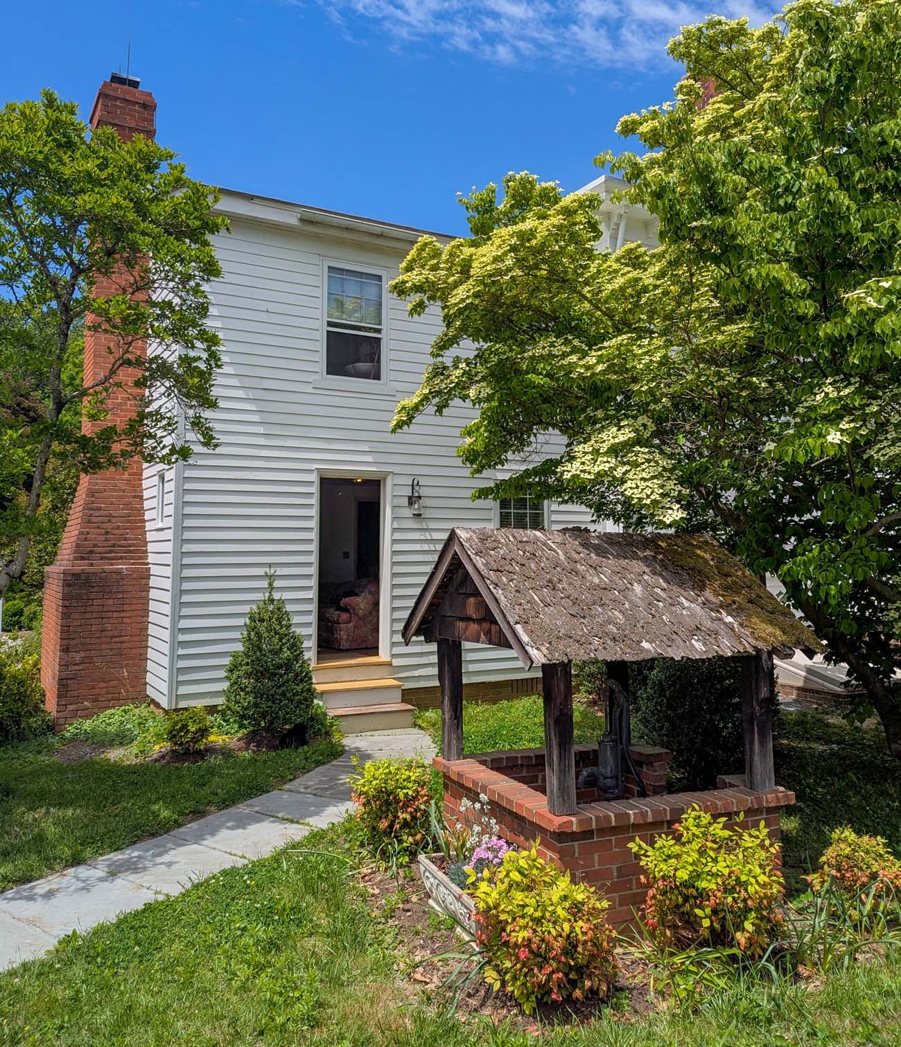 The exterior of the Fairy Hill Suite at Brampton 1860 in Chestertown, Maryland, a white clapboard two-story structure with a red brick chimney, surrounded by spring greenery including a blooming dogwood tree, with the property's original moss-covered brick and timber well structure prominently in the foreground