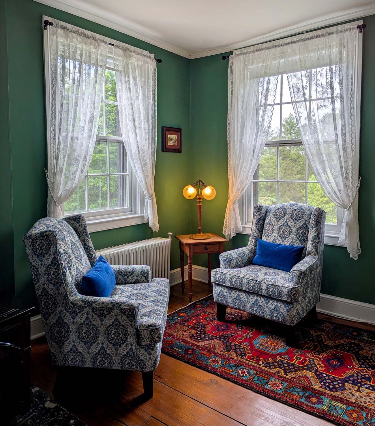 A sitting area inside the Brampton 1860 Manor House featuring deep green painted walls, two blue and white upholstered wingback chairs facing each other on a colorful vintage Persian rug, a small antique side table with a vintage amber glass lamp, and two large windows with sheer lace curtains overlooking green spring trees