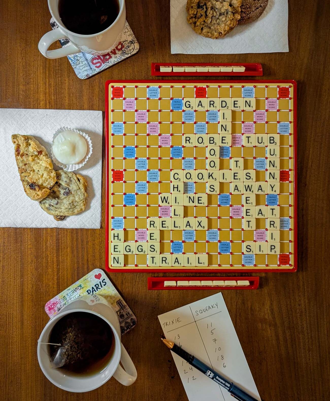 A flat-lay overhead shot of a Scrabble board on a wooden library table at Brampton 1860, with words spelled out including GARDEN, COOKIES, WINE, RELAX, EGGS, and TRAIL, alongside two cups of tea, an assortment of fresh-baked cookies and scones on napkins, and a handwritten score sheet showing Trixie and Squeaky as the players