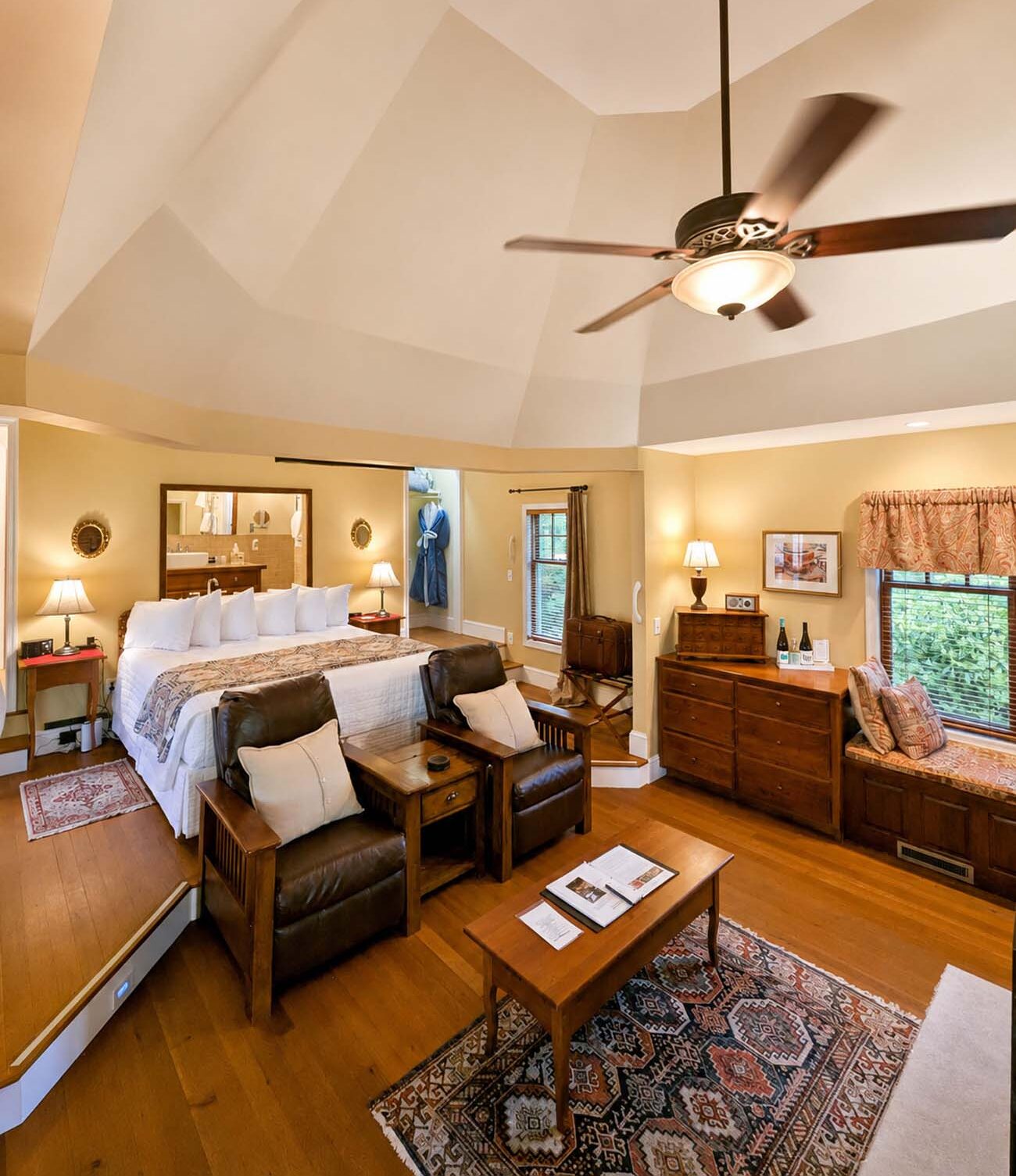 The interior of Mulberry Cottage at Brampton 1860, a spacious room with a dramatic octagonal vaulted white ceiling, a ceiling fan, a king bed with white quilted linens, two leather Mission-style recliner chairs with cream pillows flanking a coffee table, dark hardwood floors with a Persian rug, a large wooden dresser, and a window seat with garden views