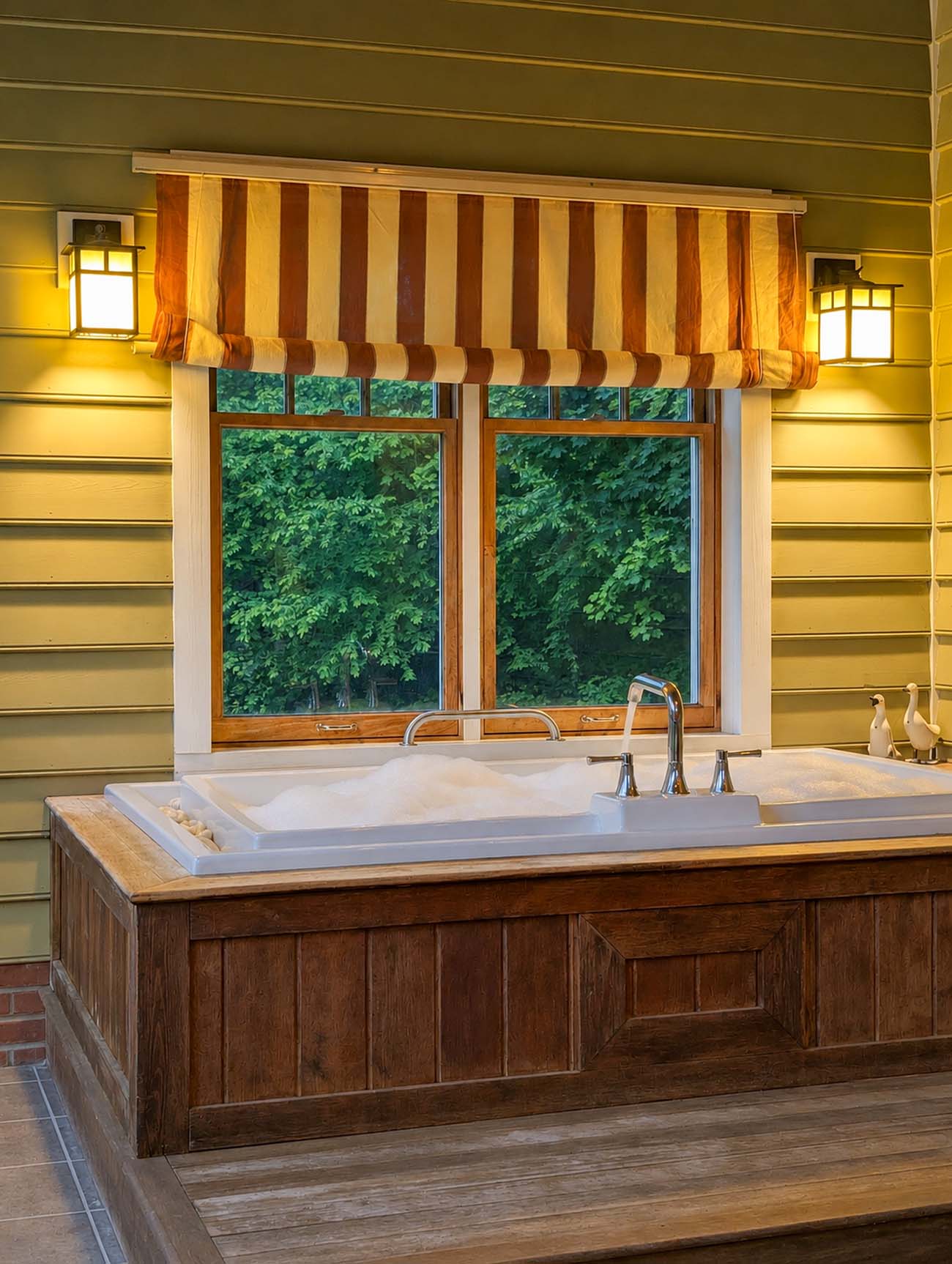 A deep rectangular soaking tub set into a dark wood-paneled platform inside Mulberry Cottage at Brampton 1860, framed by a double wooden window with a striped rust and cream Roman shade, flanked by two mission-style wall sconces casting warm light, with a view of dense green spring foliage outside the window