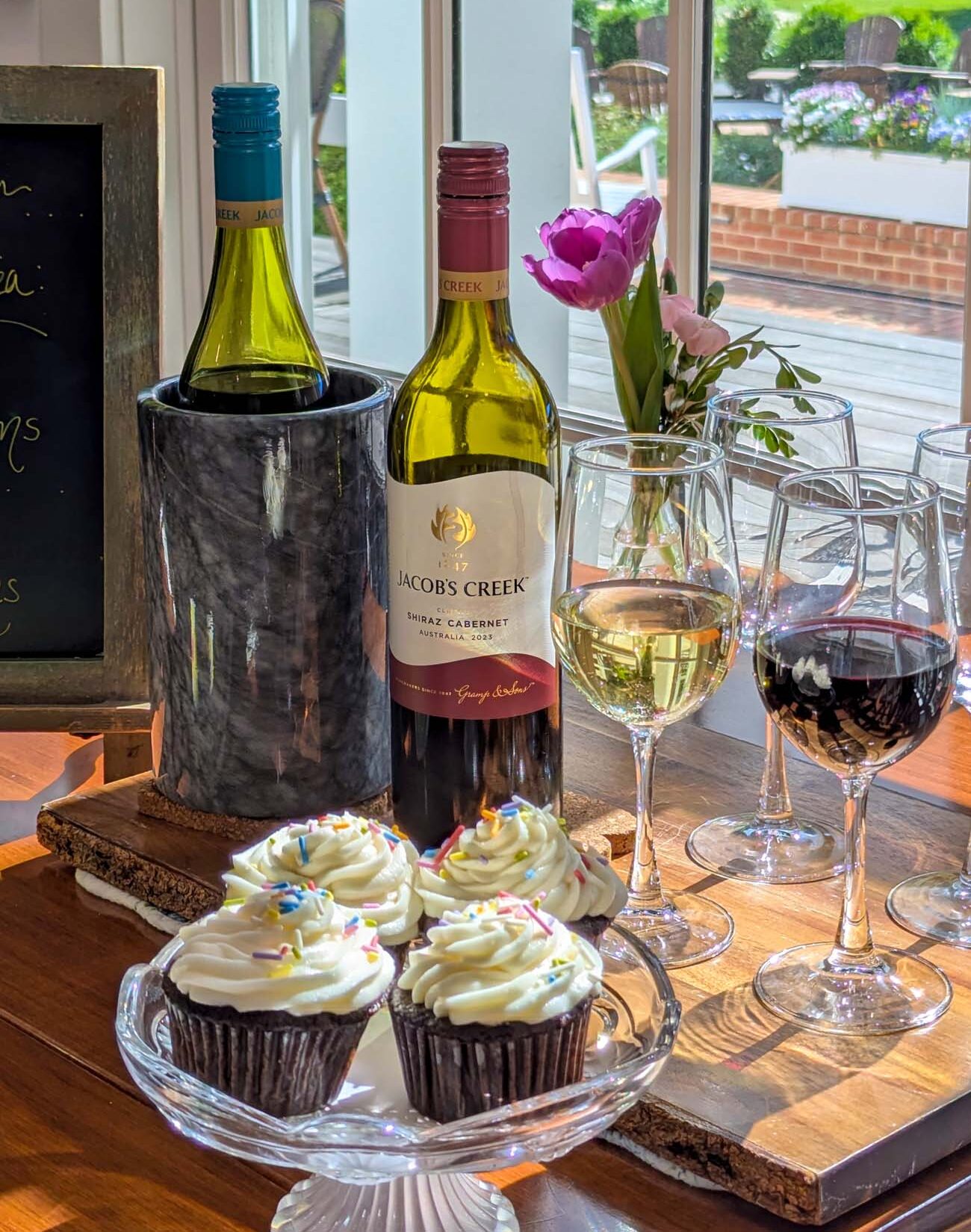 The daily tea time and social hour spread at Brampton 1860, featuring a bottle of Jacob's Creek Shiraz Cabernet and a chilled white wine in a marble wine cooler, four wine glasses with red and white wine poured, a glass cake stand holding four chocolate cupcakes with white frosting and rainbow sprinkles, a single pink tulip in a small vase, and a chalkboard menu in the background, with the Brampton porch and spring gardens visible through the window