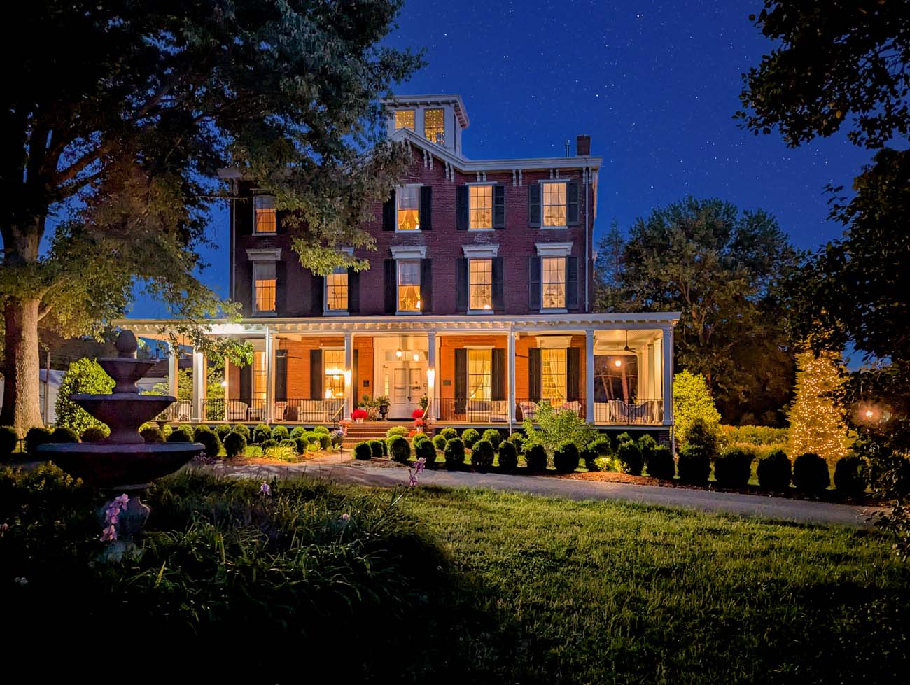 The Brampton 1860 three-story red brick historic manor house photographed at night against a deep blue star-filled sky, with warm light glowing from every window, the white columned wraparound porch illuminated, a stone fountain on the left side of the lawn lit from below, and rows of trimmed boxwood hedges lining the gravel driveway in the foreground