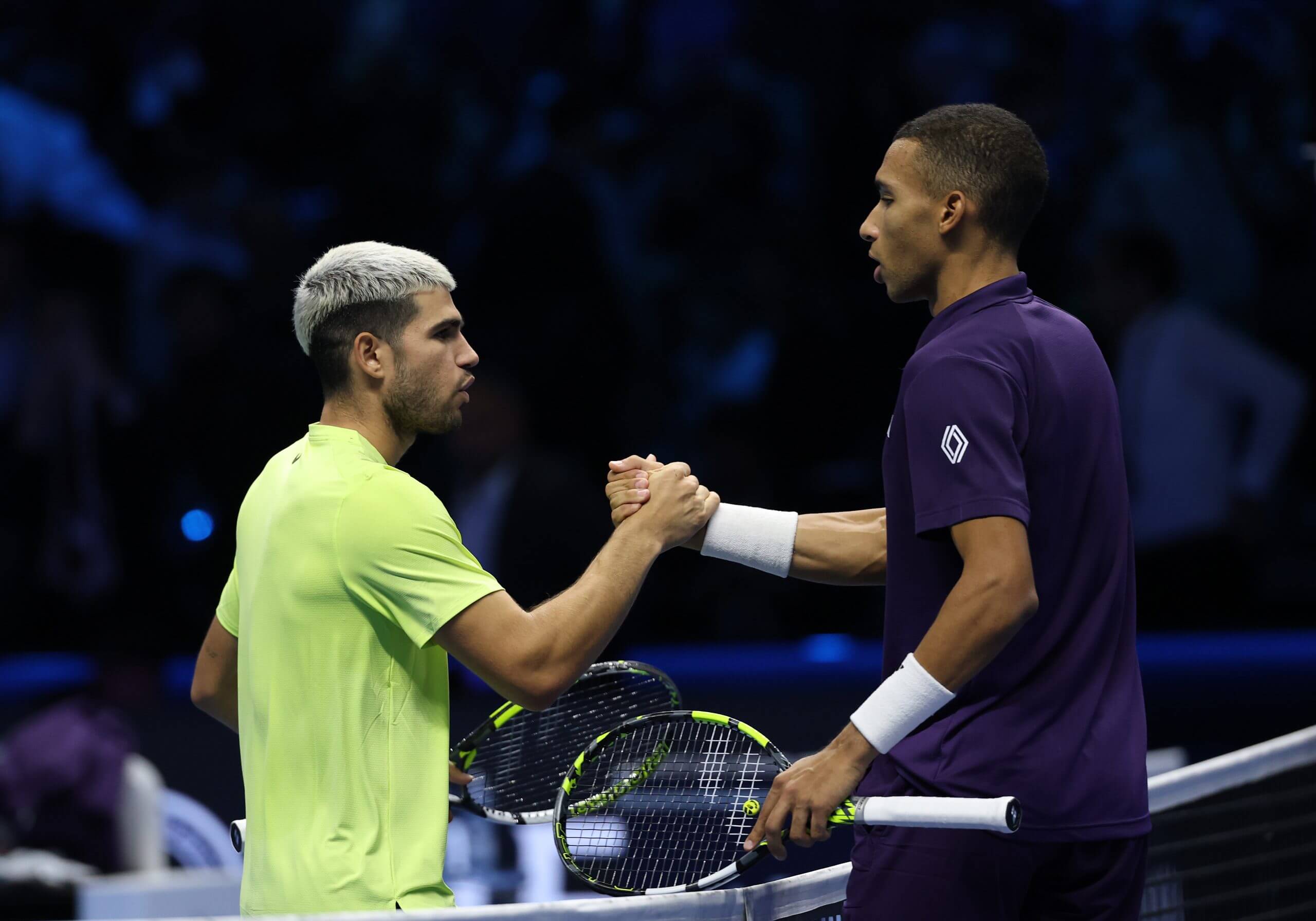 Carlos Alcaraz (left) wears a green-yellow tennis outfit as he shakes hands with Félix Auger-Aliassime (right) who wears a purple polo.