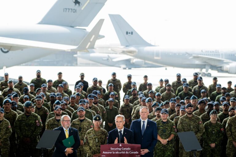 Mark Carney stands at a podium, speaking. Behind him are dozens of people wearing CAF uniforms. In the background, two planes are visible.