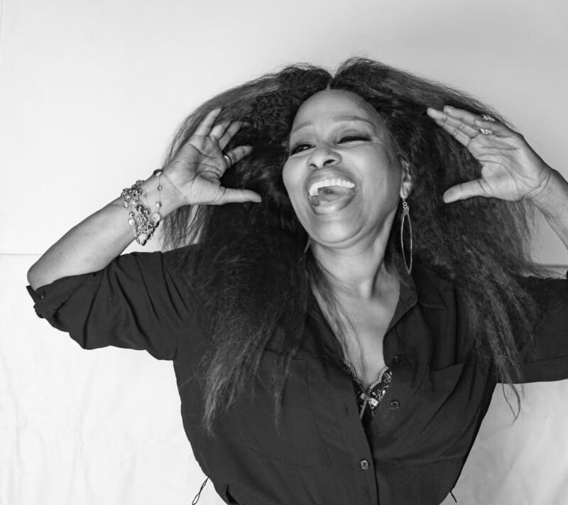 Black and white photo of a woman smiling joyfully, tilting her head back and holding her hands up by her hair. She wears hoop earrings, bracelets, and a dark shirt with voluminous curly hair.