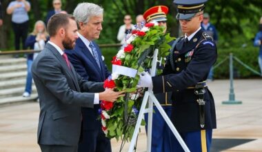 Two men in suits place a red-white-blue floral wreath at a memorial as a uniformed soldier stands at attention nearby, with onlookers in the background.