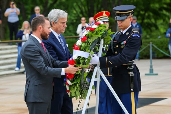 Two men in suits place a red-white-blue floral wreath at a memorial as a uniformed soldier stands at attention nearby, with onlookers in the background.