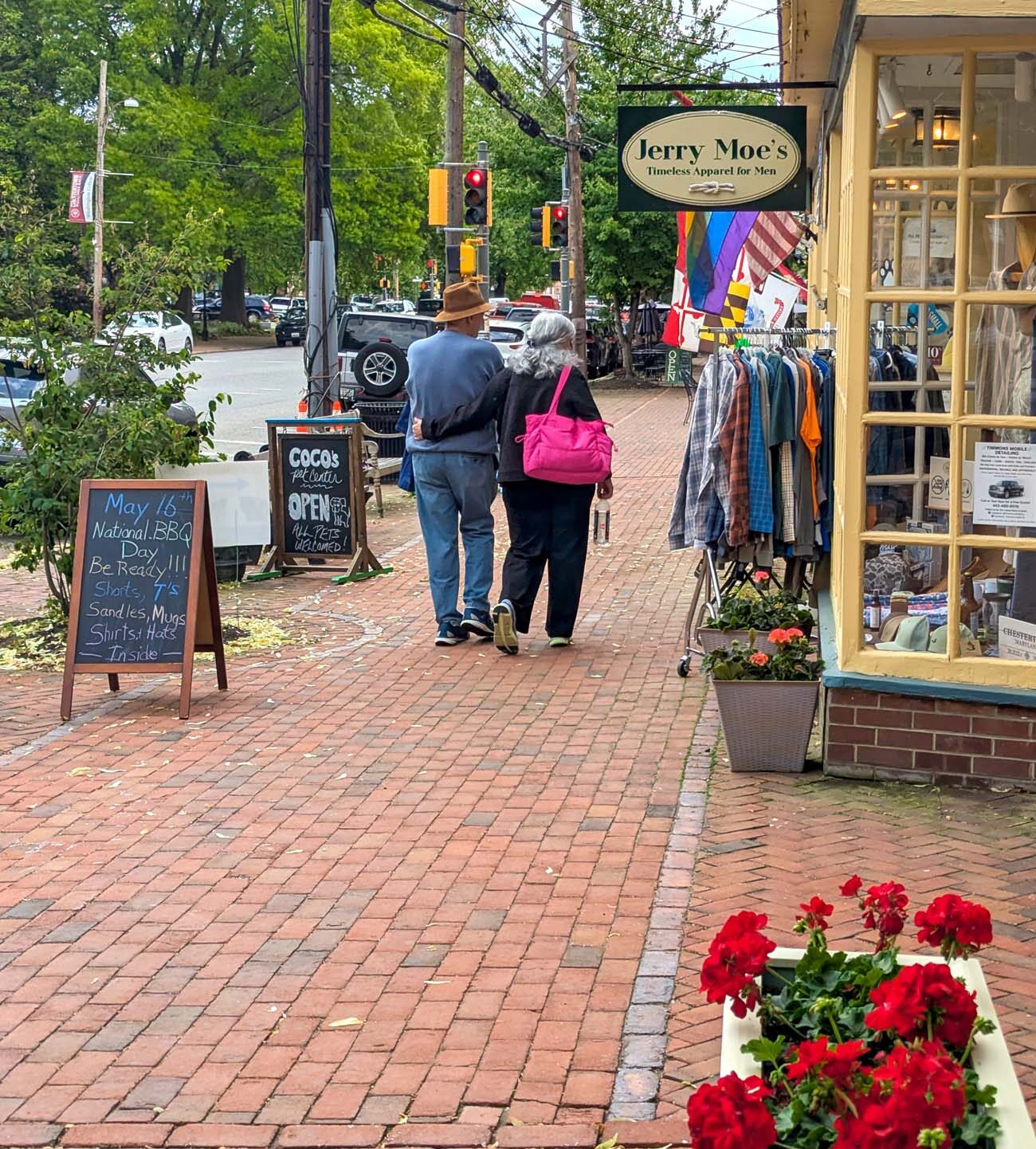 A couple walking arm in arm away from the camera along the red brick sidewalk of historic High Street in Chestertown, Maryland in spring, the man wearing a brown hat and blue jeans, the woman carrying a bright pink bag, passing the Jerry Moe's Timeless Apparel for Men storefront with clothing displayed outside, a Coco's Pet Center chalkboard sign advertising National BBQ Day on May 16th, a window box of red geraniums in the foreground, and mature green trees lining the street in the background