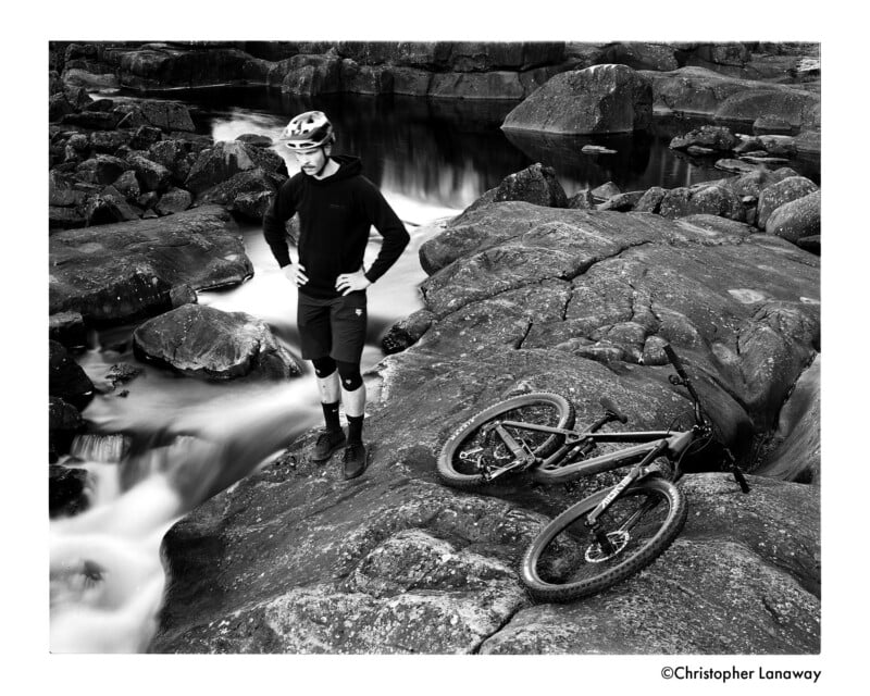 A person in cycling gear stands on rocky terrain beside a mountain bike, near a flowing stream and large boulders. The scene is in black and white, creating a dramatic and adventurous mood.