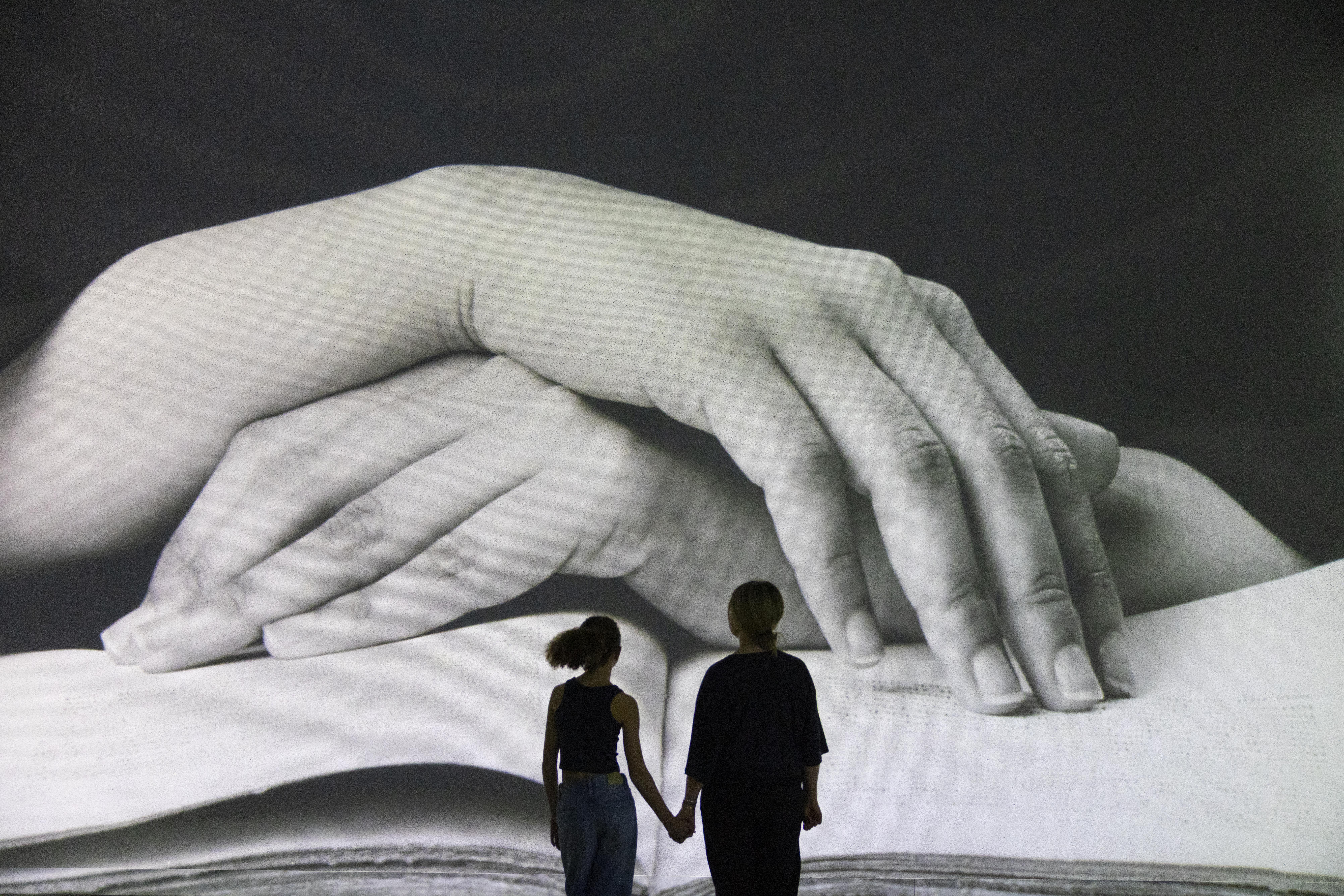 Two people stand hand in hand, seen from behind, looking up at a large-scale black-and-white projection of two hands resting on an open book.