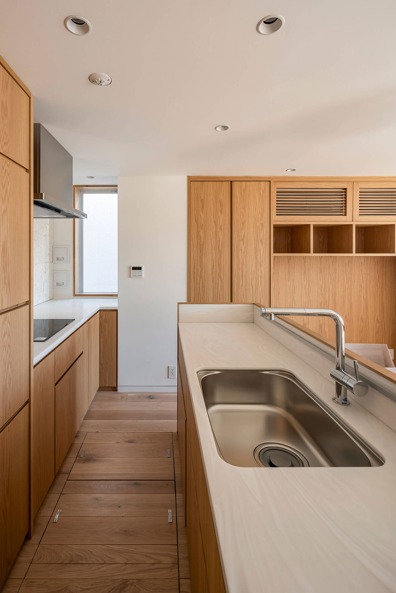 Modern kitchen with light wood cabinets, built-in appliances, and a stainless steel sink on a white countertop; natural light enters from a window.